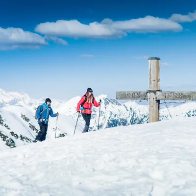 Two hikers trekking through snowy alpine landscape near summit cross